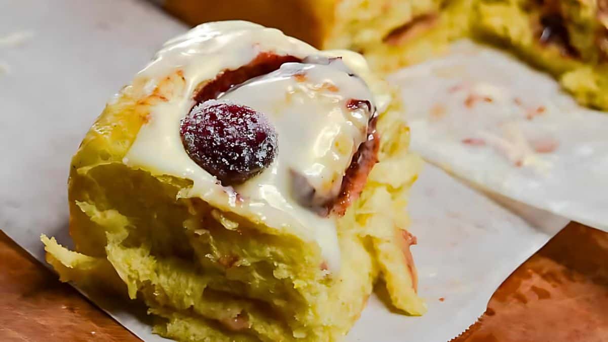 A close-up of a cinnamon roll topped with creamy icing and a single cherry. The roll is placed on a piece of parchment paper, with another partially viewed roll in the background. The icing appears thick and glossy.