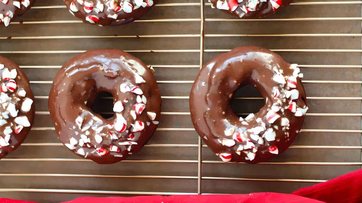 Chocolate-glazed donuts with crushed peppermint candies on top are placed on a cooling rack. A red cloth is partially visible at the bottom.