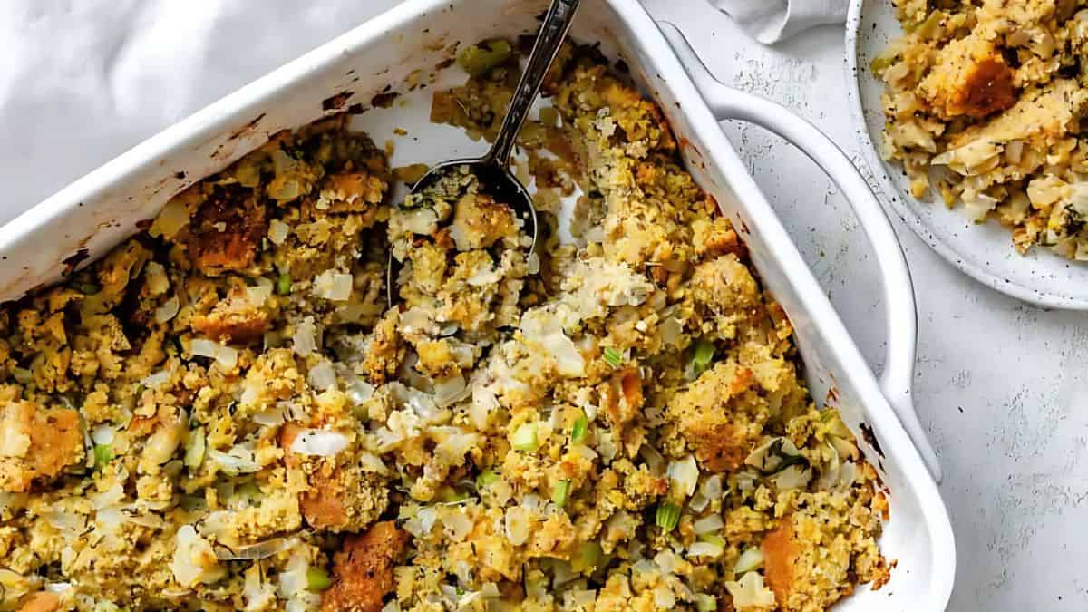 A white casserole dish filled with baked stuffing, featuring chunks of bread, onions, and herbs. A serving spoon rests inside the dish. A portion of the stuffing is served on a white plate next to the casserole.