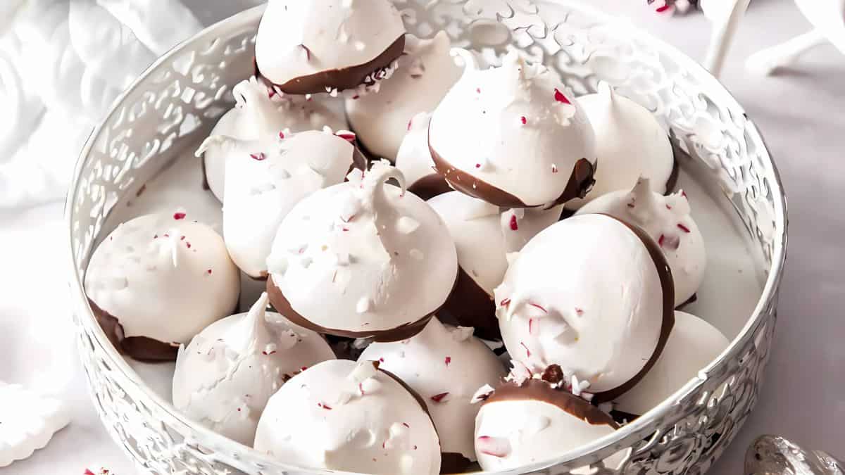 A decorative silver basket filled with white meringue cookies dipped in chocolate and sprinkled with crushed peppermint candies, set on a white surface.