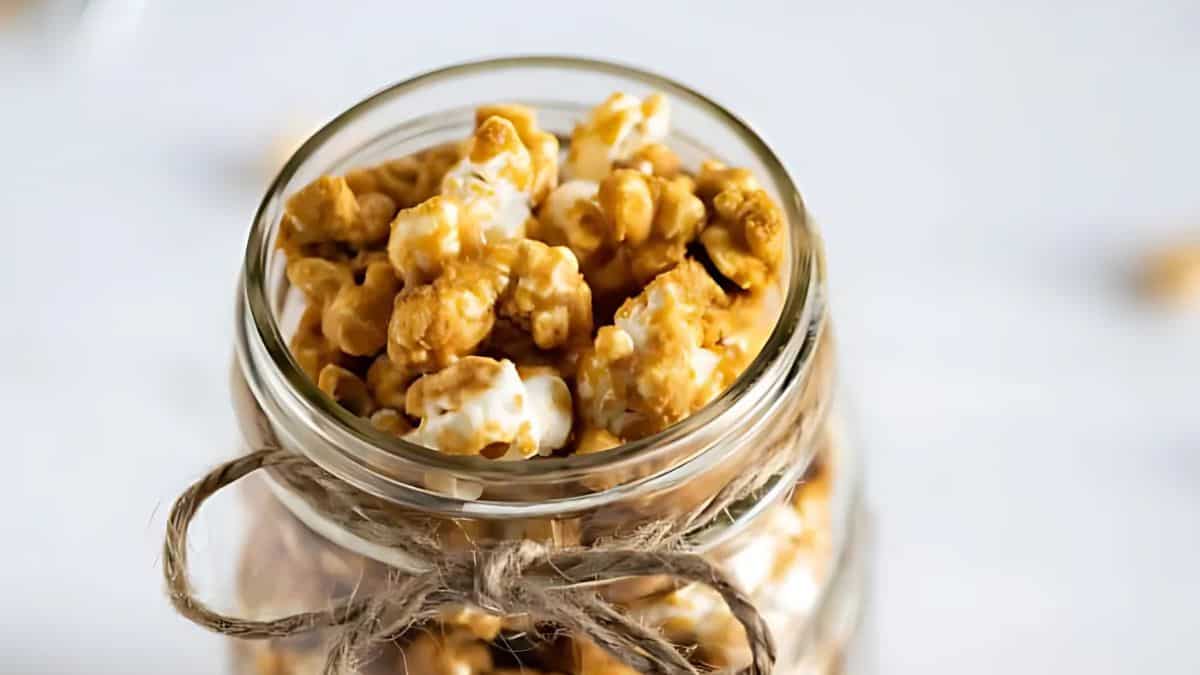 A close-up of a glass jar filled with caramel popcorn, tied with a rustic twine bow. The background is softly blurred, highlighting the golden-brown color and glistening texture of the popcorn.