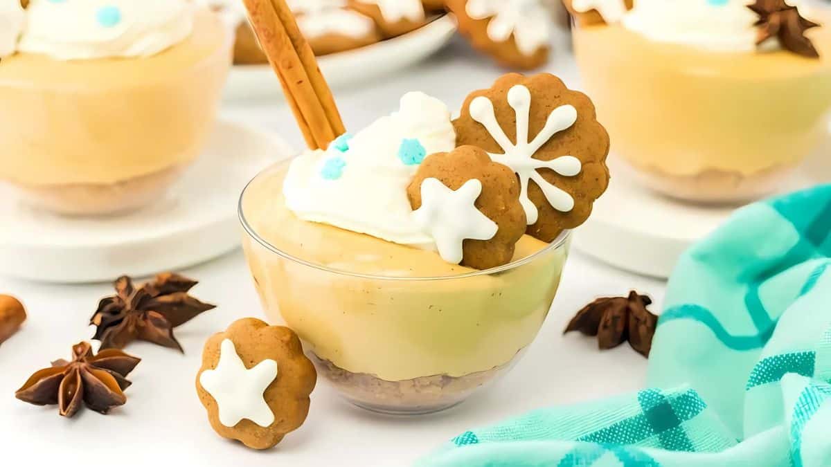 A glass bowl filled with creamy dessert, topped with whipped cream, a cinnamon stick, and star-shaped gingerbread cookies with white icing. Star anise and more dessert dishes are in the background. A teal cloth is on the side.
