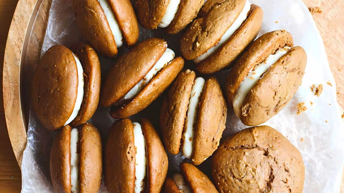 A plate of brown sandwich cookies filled with white cream is displayed. The cookies are unevenly spread across a crumpled sheet of wax paper, showcasing their inviting texture and rich filling.
