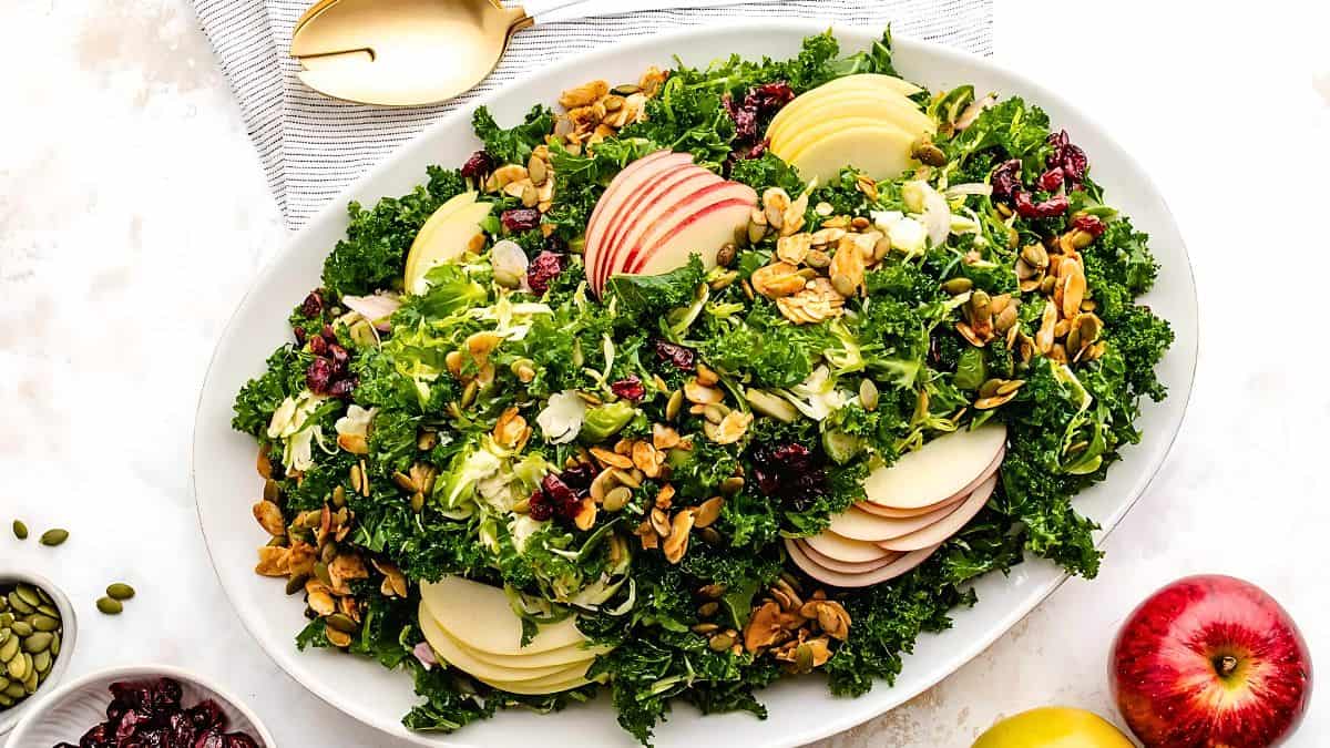 A plate of salad featuring curly kale, apple slices, dried cranberries, pumpkin seeds, and nuts. A gold-colored fork and spoon rest beside the plate, and apples are placed nearby on a light surface.