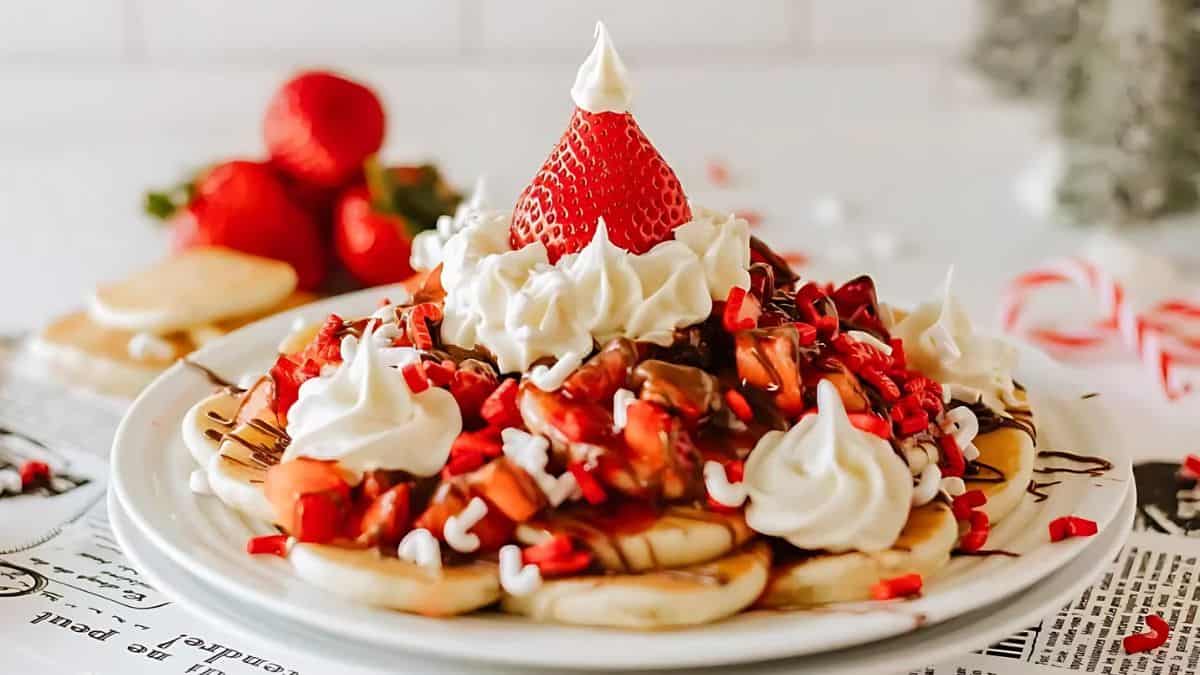 A festive dessert featuring a plate of whipped cream-topped pancakes garnished with sliced strawberries and chocolate syrup. The centerpiece is a strawberry shaped like a Santa hat, surrounded by red and white candy pieces.