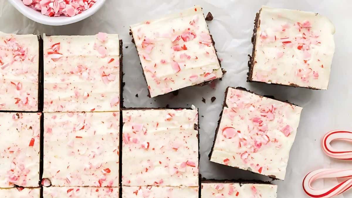 A batch of chocolate peppermint bark, cut into squares, is displayed on a white surface. Each piece is topped with a layer of white chocolate and sprinkled with crushed peppermint candy. A bowl of crushed candy and a few candy canes are nearby.