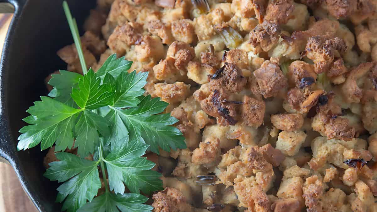 Close-up of a skillet filled with golden-brown stuffing topped with fresh green parsley leaves. The stuffing appears crispy with chunks of toasted bread and herbs.