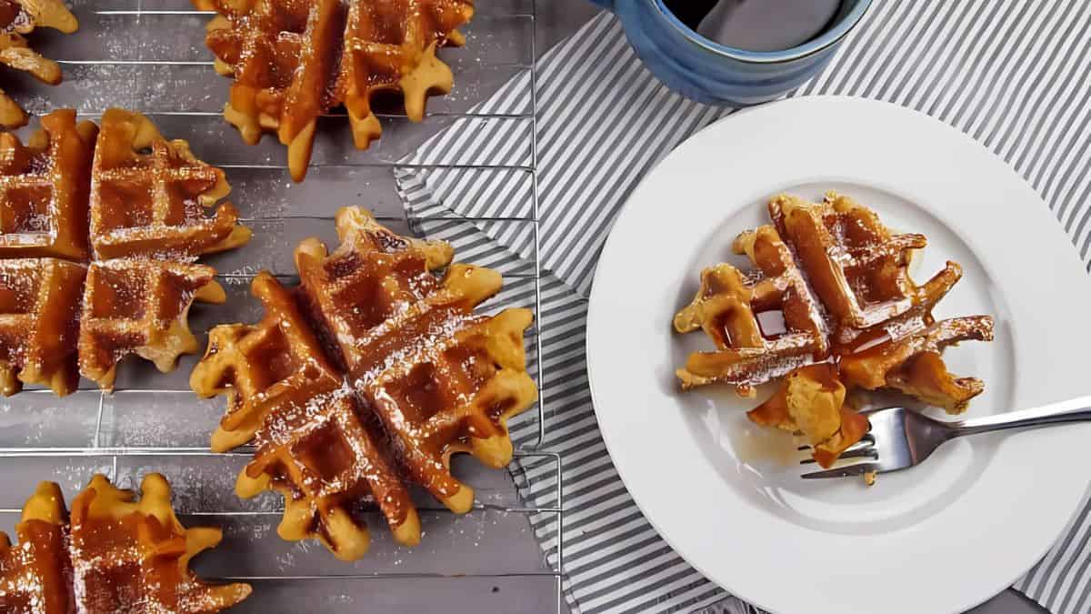 Golden waffles on a cooling rack, with one served on a white plate alongside a fork. The waffles are dusted with powdered sugar. A striped napkin and a blue cup complete the breakfast setting.