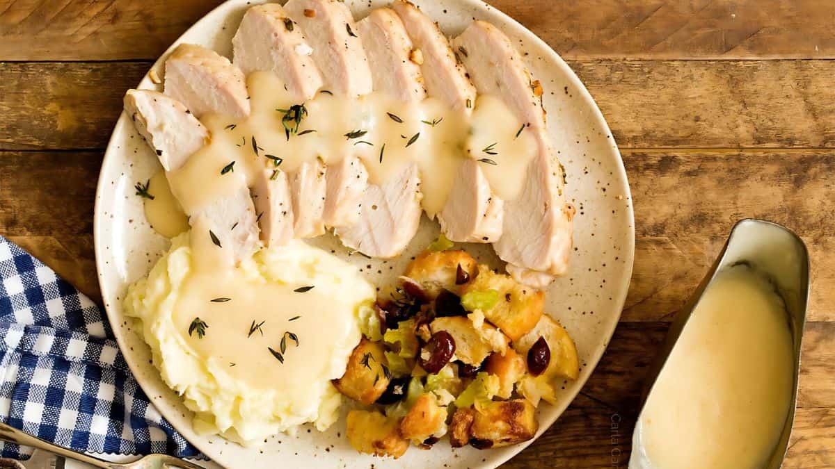 A plate with sliced turkey breast topped with gravy, served alongside mashed potatoes with gravy and stuffing with cranberries. A gravy boat is nearby, and a blue checkered napkin is on the side.