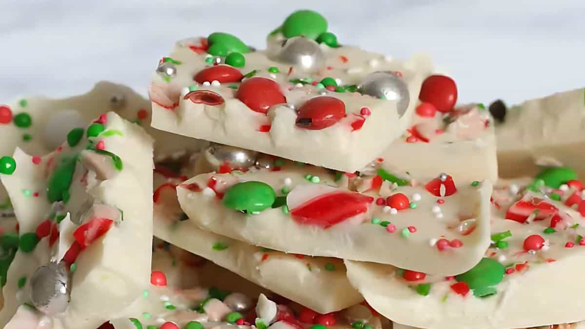 A stack of white chocolate bark pieces topped with festive red, green, and silver sprinkles, candy-coated chocolates, and crushed peppermint candies. The background is blurred, highlighting the colorful toppings on the bark.
