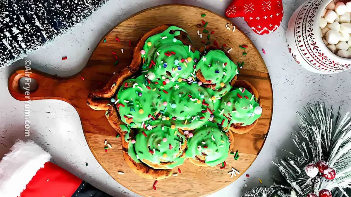 A round wooden board holds Christmas tree-shaped pastries topped with bright green icing and colorful sprinkles. Snowflake designs, a black and white stocking, a red mitten, and a bowl of marshmallows complete the festive scene.