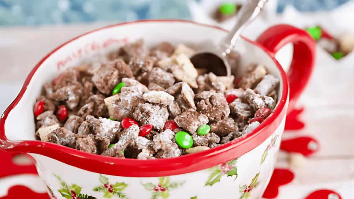 A festive bowl filled with chocolate-coated cereal snack mix, dusted with powdered sugar and interspersed with red and green candies. The bowl has a Christmas-themed holly design and a spoon rests inside.