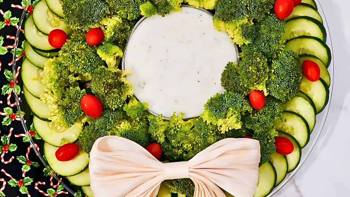 A festive holiday platter arranged like a wreath, featuring broccoli florets, cucumber slices, cherry tomatoes, and a white dip in the center. A large white bow is placed at the bottom, adding a decorative touch.