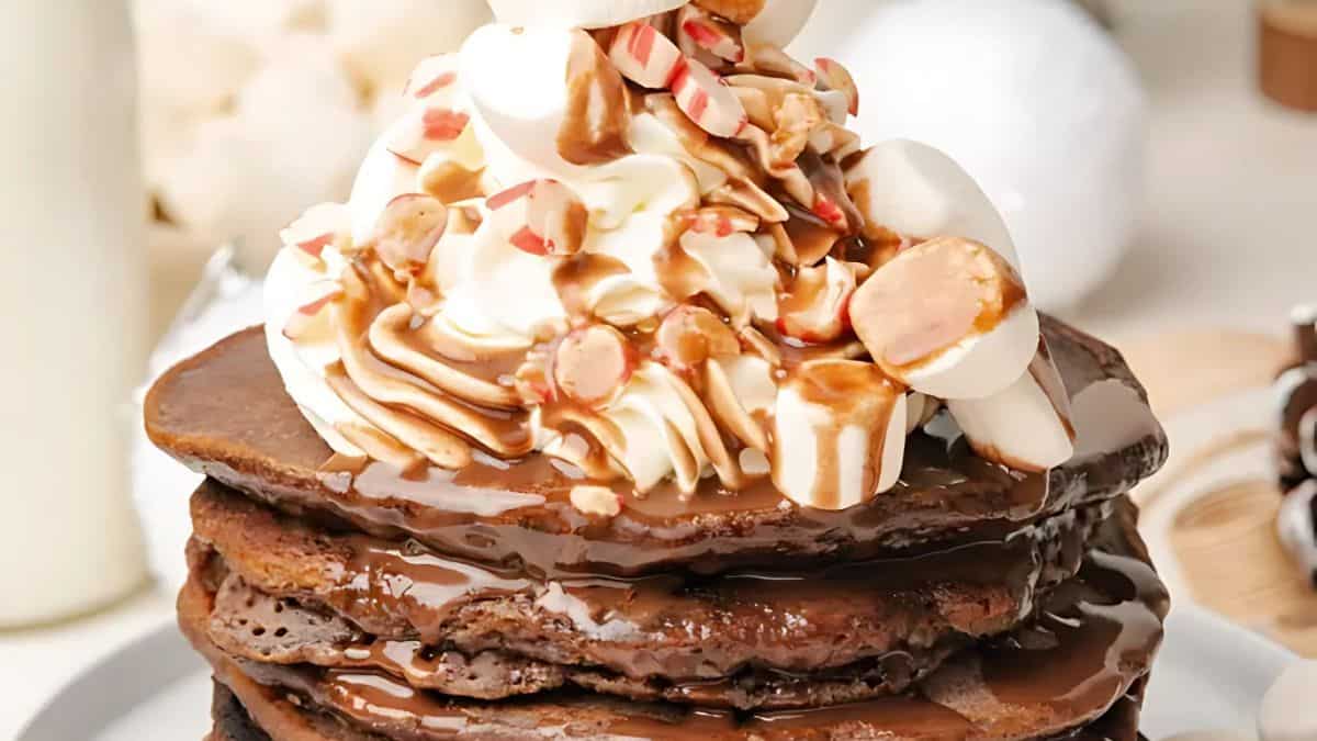 A stack of chocolate pancakes topped with whipped cream, marshmallows, caramel sauce, and crushed peppermint candies on a plate.