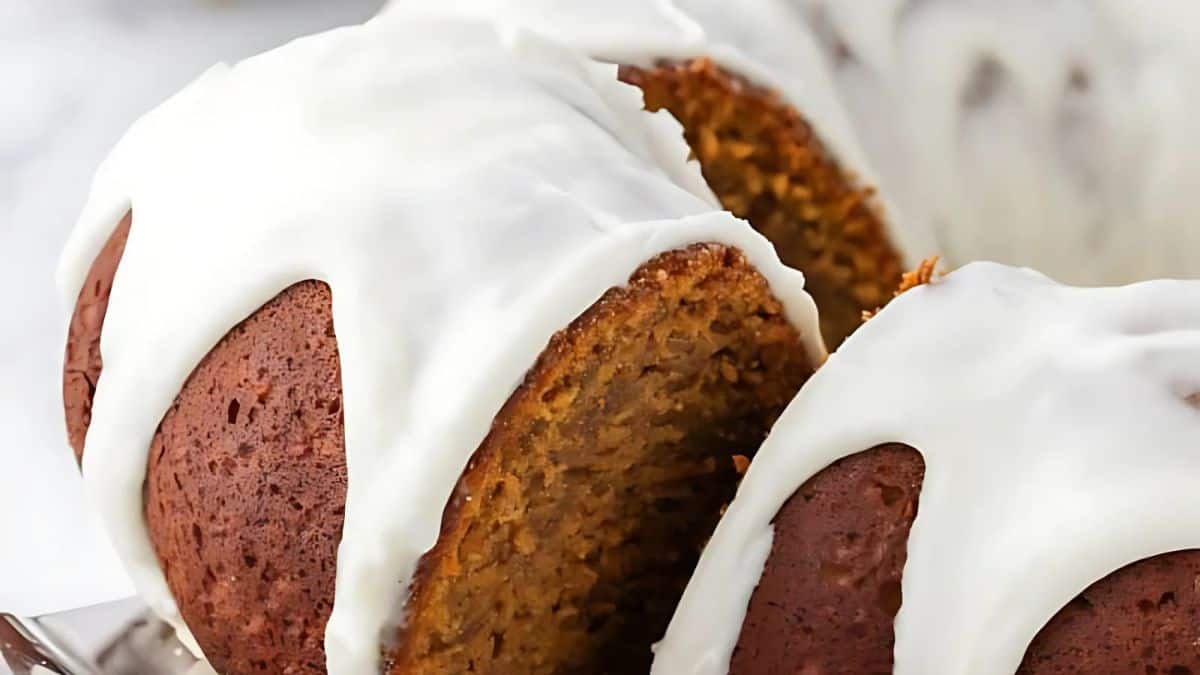 A close-up of a sliced pumpkin bundt cake topped with a thick layer of white icing. The cake's moist texture and autumnal orange color are visible. The background is blurred, highlighting the cake's rich details.