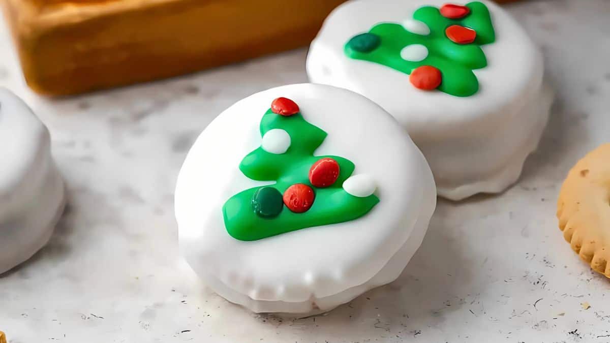 A close-up of a cookie covered in white icing, decorated with a green Christmas tree design and red and white candies as ornaments. Another cookie is partially visible in the corner. The surface is a light, textured background.