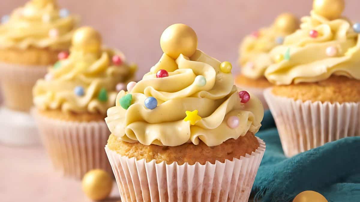 Cupcakes with creamy swirls of frosting topped with colorful sugar pearls and gold spheres. The cupcakes are displayed in white paper wrappers on a table with a teal cloth.