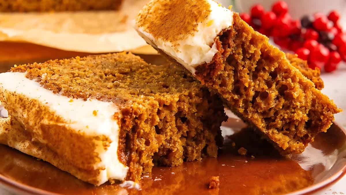 Two slices of fluffy gingerbread cake topped with creamy icing are arranged on a wooden plate. Red berries are blurred in the background, adding a festive touch.