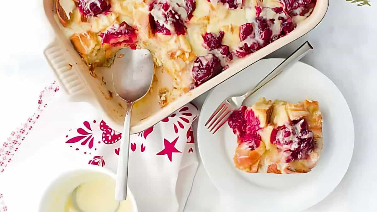 A casserole dish of bread pudding topped with cherries and a creamy sauce sits next to a white plate with a serving and a fork. A silver serving spoon rests in the dish. A bowl of extra sauce is nearby on a decorative napkin.