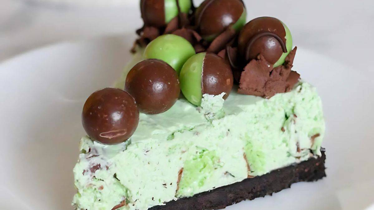 A slice of mint chocolate dessert topped with green and brown chocolate balls on a plate. The dessert has a green mint-flavored creamy layer with chocolate shavings, set on a dark chocolate crust.