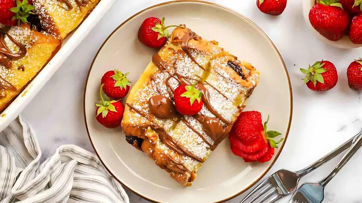 A slice of dessert topped with powdered sugar and chocolate drizzle is served on a plate with fresh strawberries. The plate is accompanied by a fork and knife on a striped napkin. Nearby, more strawberries and a baking dish are visible.