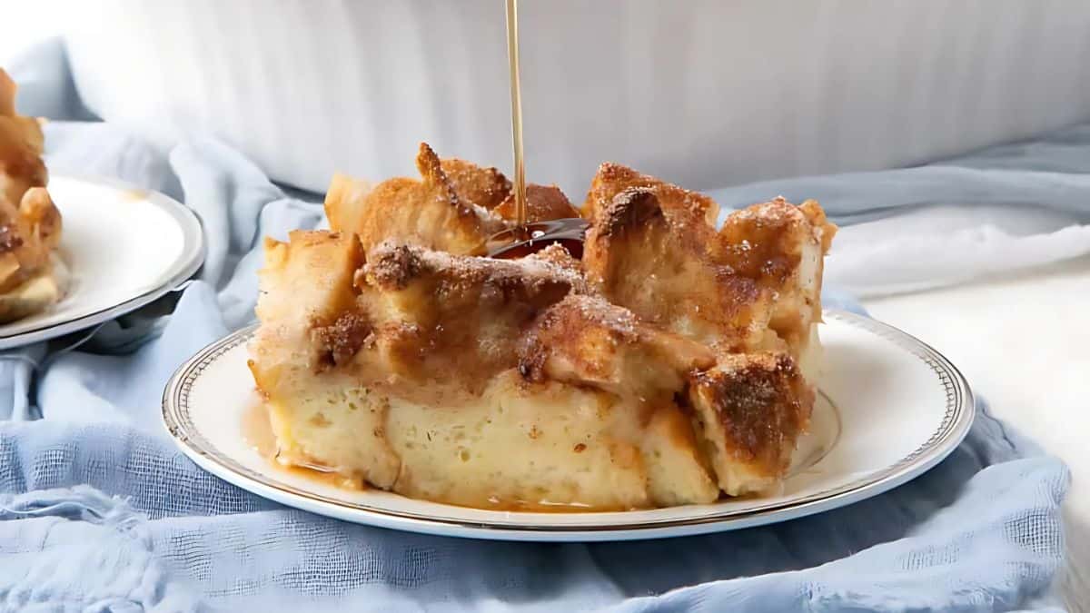 A slice of bread pudding drizzled with syrup on a white plate. The dessert is golden brown, with a slightly crisp texture on top, set against a light blue fabric backdrop.