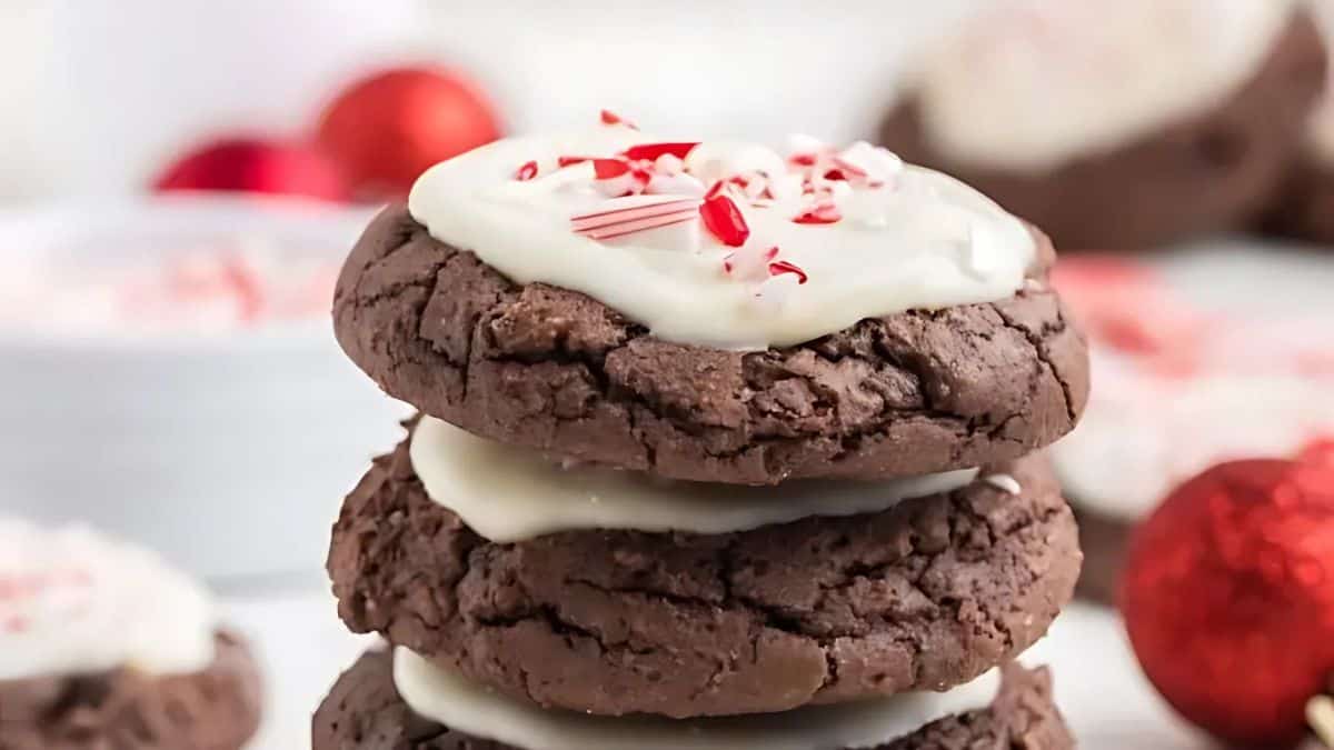 A stack of three chocolate cookies topped with white icing and crushed peppermint candy pieces. The background features blurred cookies and red decorative ornaments.