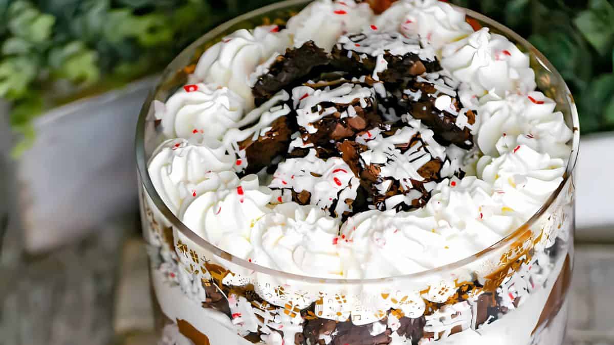 A layered dessert in a glass bowl, featuring whipped cream, chocolate pieces, and drizzled chocolate sauce. Peppermint candy bits are scattered on top. The background shows green foliage out of focus.