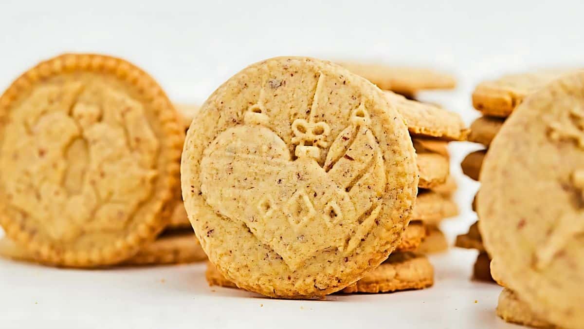 Close-up of round, decorative cookies with a carved heart and arrow design. The cookies are stacked unevenly in the background on a white surface.