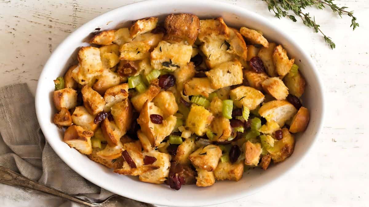 A white oval dish filled with homemade stuffing, featuring golden-brown bread cubes, celery, and cranberries. A sprig of fresh thyme lies beside the dish on a white wooden surface.