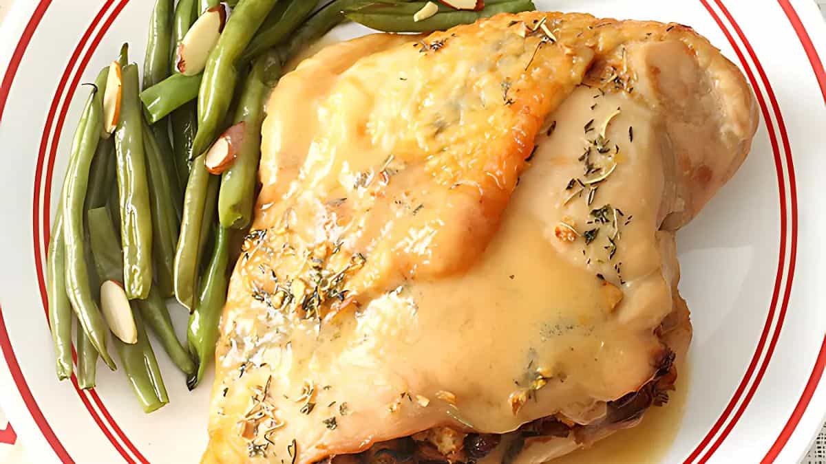 A plate with roasted chicken covered in gravy, garnished with herbs, next to a serving of green beans sprinkled with almond slices. The dish is presented on a white plate with a red border.