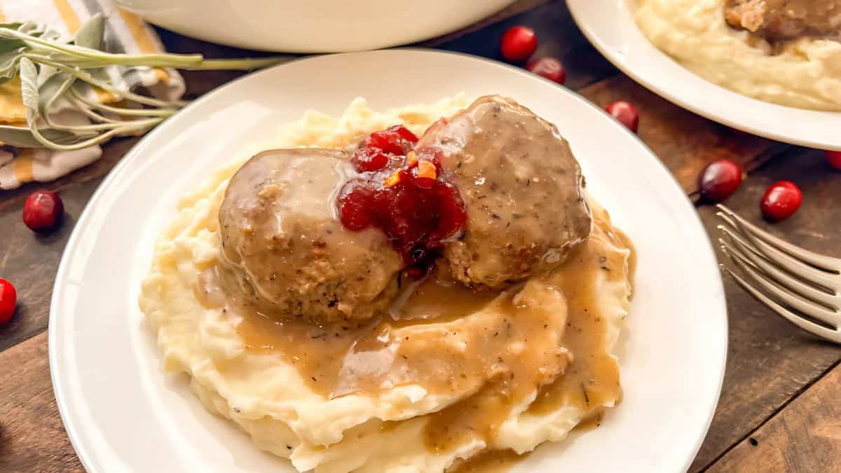 A plate with mashed potatoes topped with two meatballs, gravy, and cranberry sauce. The plate is surrounded by scattered cranberries on a wooden table, with a fork and a cloth napkin nearby.