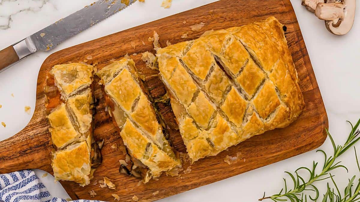 A golden-brown puff pastry on a wooden board, partially sliced to reveal a filling of mushrooms and herbs. A knife lies nearby, with rosemary and a mushroom on the right, and a blue striped cloth on the left.