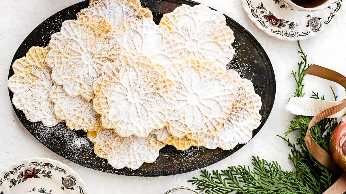 A black plate with several thin, intricately patterned pizzelle cookies dusted with powdered sugar. The table is set with teacups, a sprig of pine, and a ribbon, creating a festive ambiance.