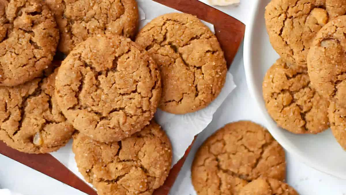 A close-up of a batch of freshly baked peanut butter cookies on a wooden board and a white plate. The cookies have a golden-brown color with a cracked surface, creating an inviting and homemade appearance.