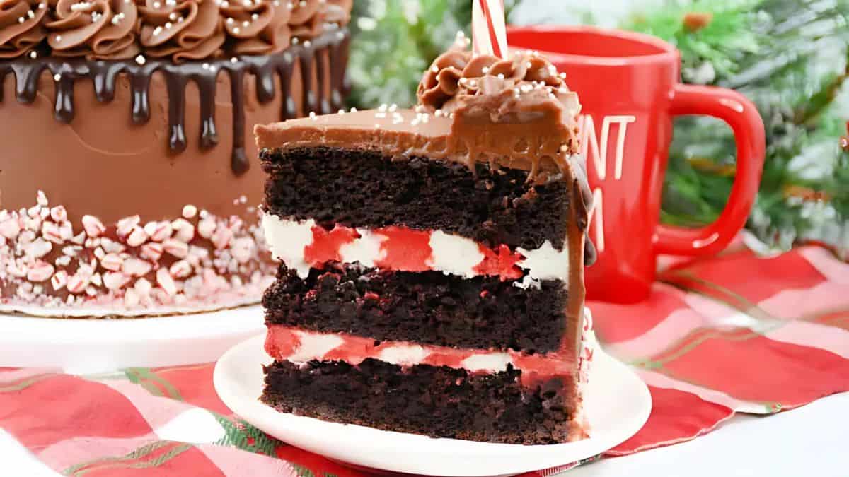 A slice of layered chocolate cake with red and white frosting, topped with chocolate ganache and a peppermint stick, sits on a white plate. A red mug and festive decorations are in the background.