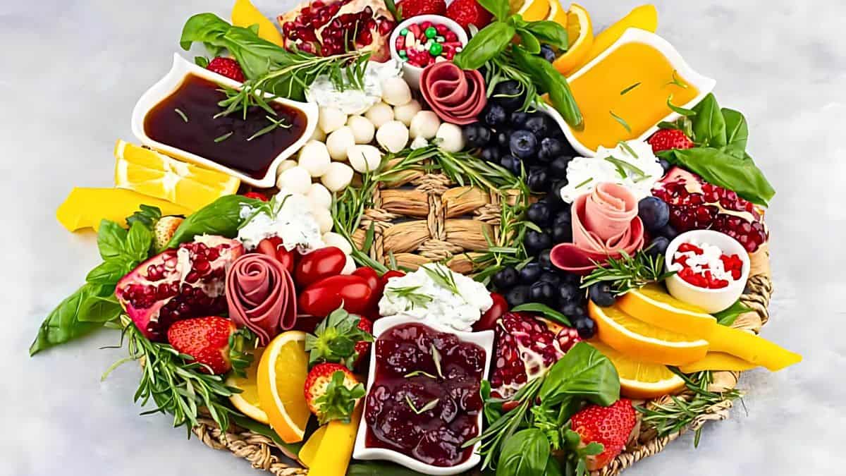 A colorful charcuterie board arranged in a circular wreath shape. It features assorted cheeses, sliced meats, olives, strawberries, blueberries, pomegranate seeds, orange slices, bell pepper strips, and fresh herbs on a woven placemat.