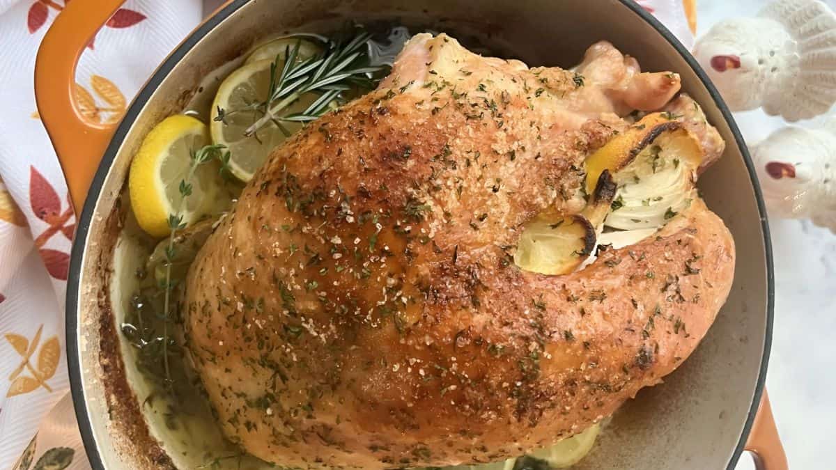 A seasoned and roasted turkey breast in a pot, garnished with slices of lemon and sprigs of rosemary. The dish is surrounded by festive decor with hints of autumn leaves.