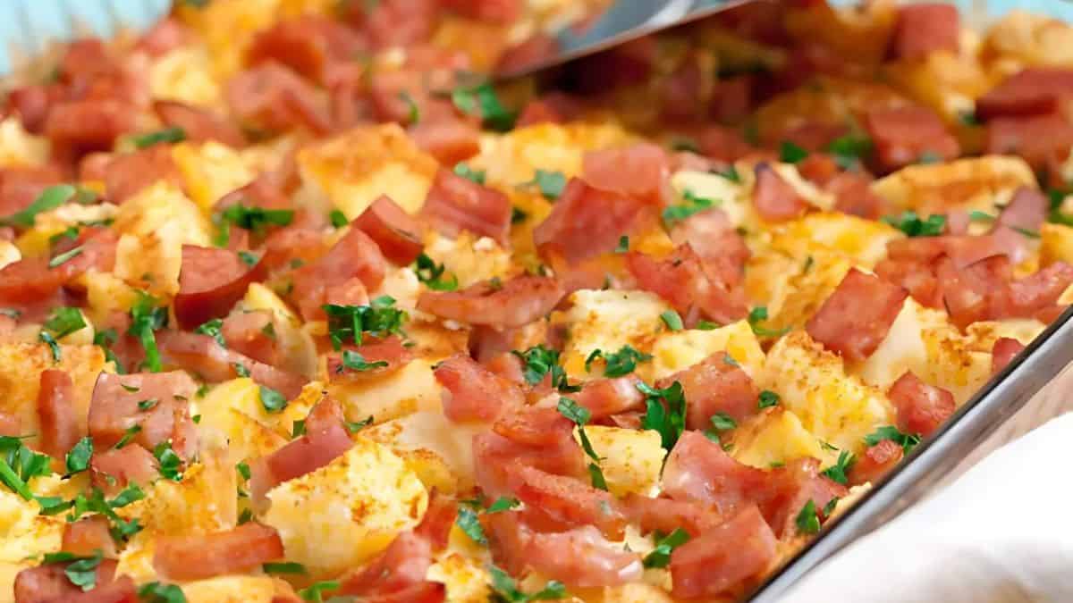 A close-up of a baked dish in a pan, featuring diced potatoes, cubes of ham, and chopped fresh parsley on top. The food appears golden brown and is garnished with herbs, adding a touch of green.
