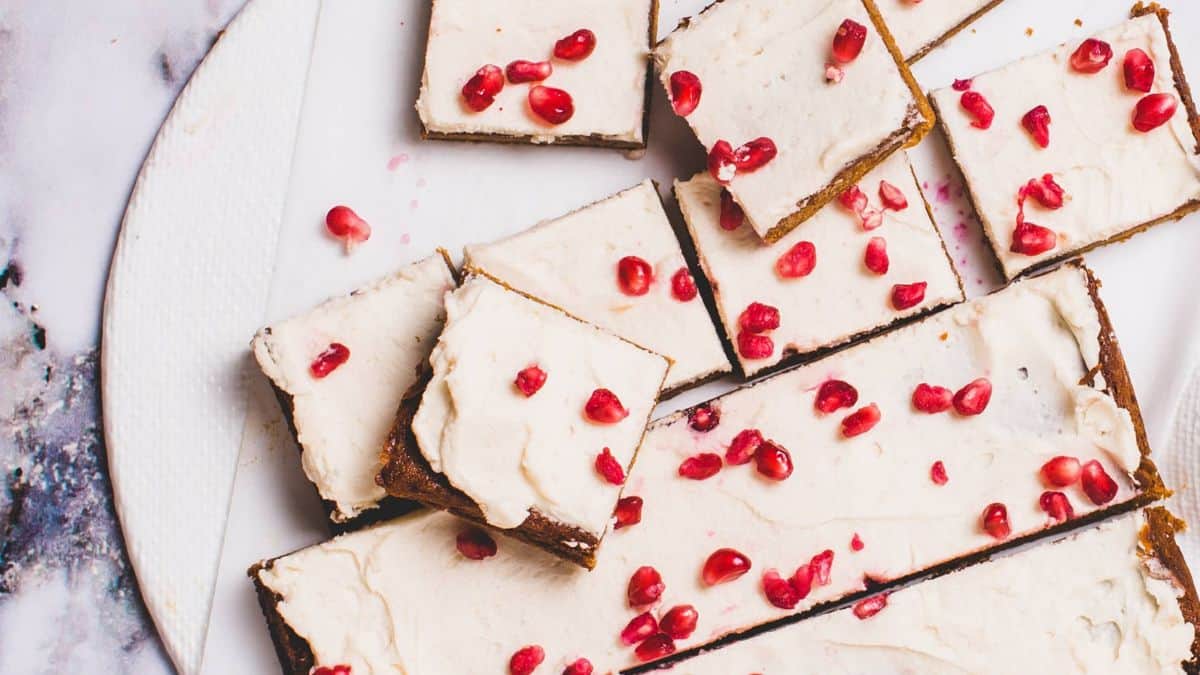 Top view of sliced dessert bars topped with white frosting and scattered pomegranate seeds on a round white plate. The background is a marble surface, creating a contrast with the vibrant red seeds.