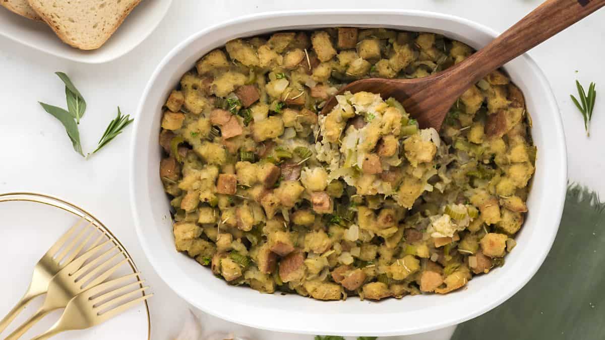 A casserole dish filled with herb stuffing with chunks of bread and vegetables. A wooden spoon rests on top. Golden forks and slices of bread are placed nearby on a white surface with green accents.