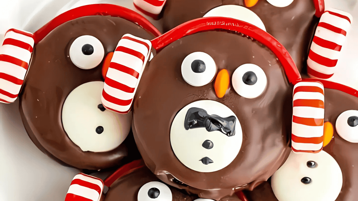 Chocolate-covered penguin-shaped cookies with candy eyes and orange beaks are decorated with red and white striped earmuffs.