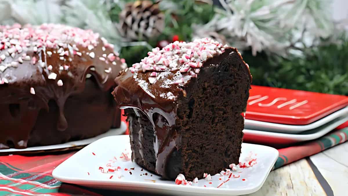 A slice of chocolate cake topped with a glossy frosting and crushed peppermint candies on a white plate. More cake is in the background, set against a festive backdrop with greenery and a stack of red plates.