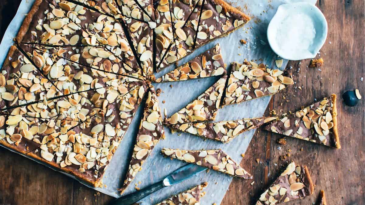 A large rectangular chocolate bark topped with sliced almonds, cut into triangular pieces, is laid out on parchment paper on a wooden table. A small bowl of coarse salt is nearby, with some pieces scattered around.