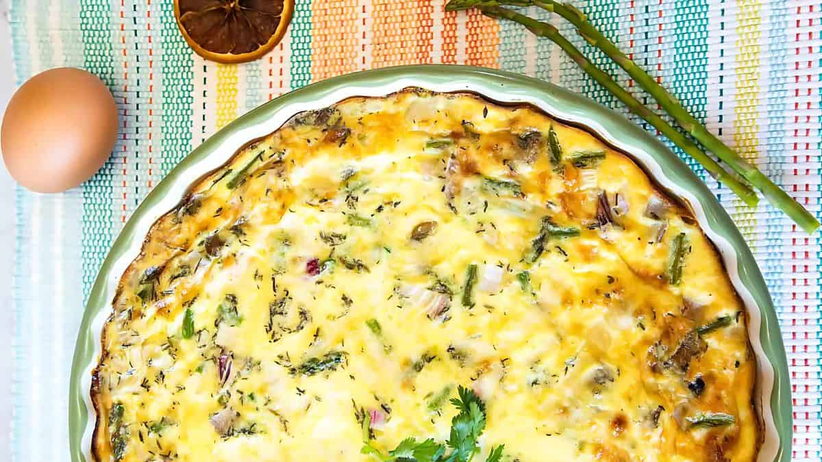 A freshly baked quiche in a round dish is placed on a colorful striped cloth. Nearby are two asparagus stalks, an egg, and a slice of dried lemon. The quiche is garnished with herbs, adding a vibrant touch to the dish.
