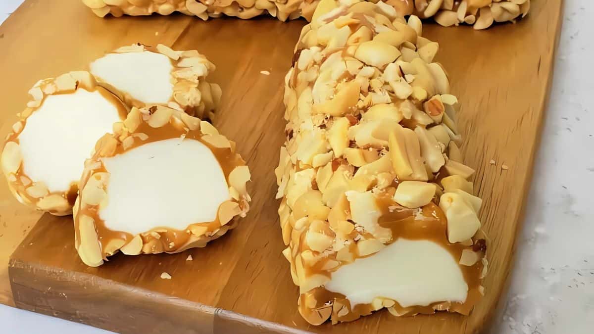 Sliced dessert log coated with crushed nuts on a wooden board. The inside is white, and the surface is covered with caramel and chopped nuts.