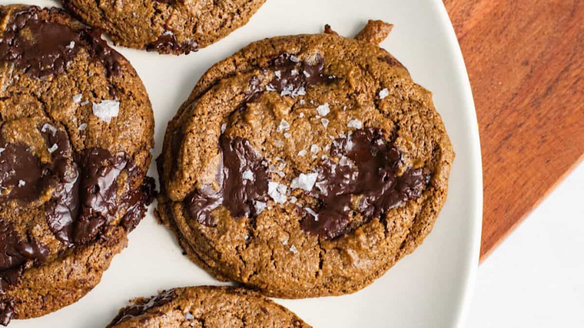 Close-up of a white plate holding four chocolate chip cookies sprinkled with sea salt flakes. The cookies are golden-brown with visible melted chocolate chunks. The plate is on a wooden surface.