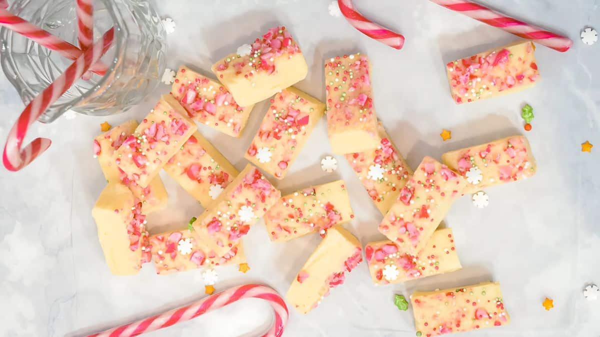 Festive holiday fudge pieces scattered on a marble surface, topped with pink sprinkles and small white and green decorations. Candy canes are placed around the fudge, adding to the festive theme.