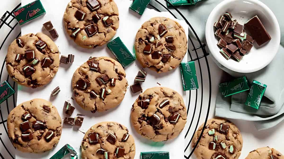 A batch of freshly baked chocolate chip cookies with chunks of mint chocolate scattered on a cooling rack. Nearby, there are unwrapped mint chocolate pieces and a bowl filled with more mint chocolates. The scene is set on a white surface.
