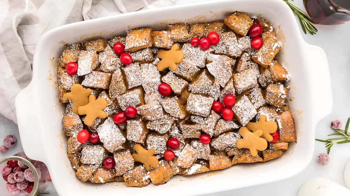 A rectangular dish filled with French toast casserole topped with powdered sugar, red cranberries, and gingerbread-shaped decorations. Surrounding the dish are sprigs of rosemary and a white cloth.